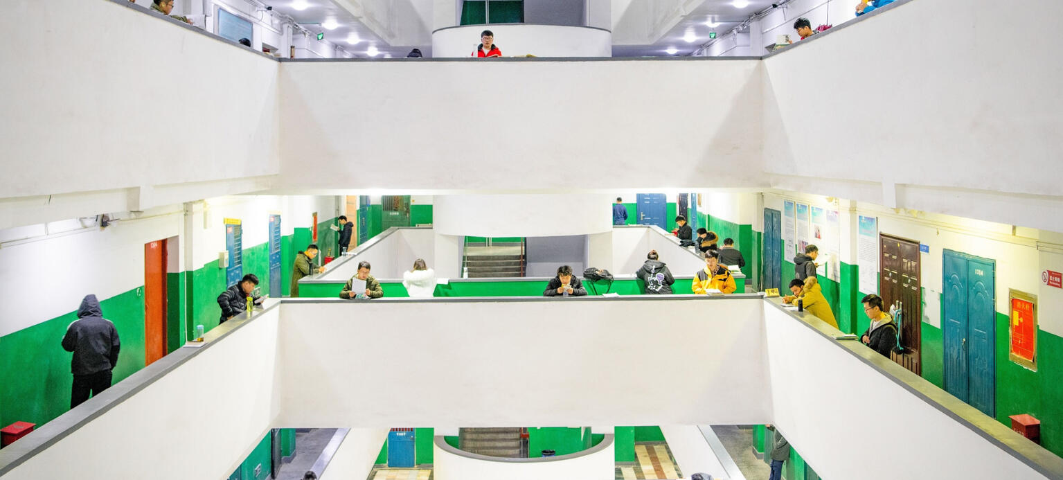 Menschen stehen auf der Galerie eines Gebäudes, das wie eine Universität wirkt und sehen über das Geländer hinunter in einen Innenhof. Viele haben Trinkflaschen und Papier dabei und scheinen zu lernen.