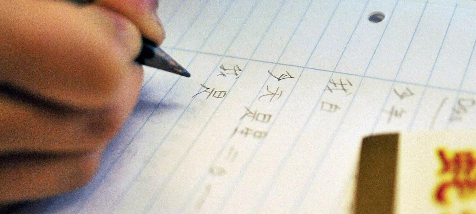 Eine nicht erkennbare Person hält einen Bleistift in der Hand und schreibt chinesische Schriftzeichen in ein Heft